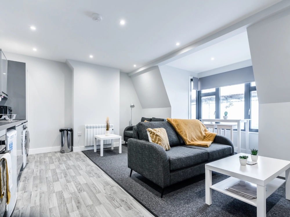 Modern open plan living room with grey sofa, yellow throw, white coffee table, grey carpet, and light wood flooring