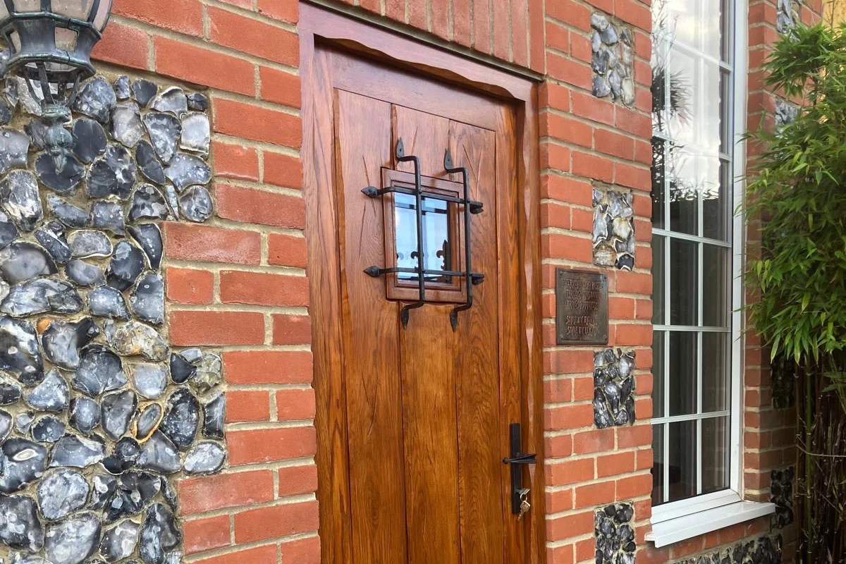 Wooden front door with black metal grille, glass panel, red brick wall, and flint exterior detail