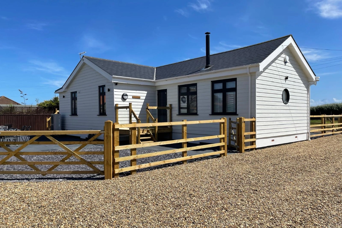 White timber clad bungalow with black framed windows, gravel driveway, wooden fencing, and blue sky